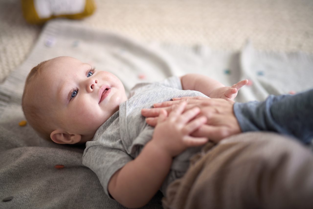 Baby liegt auf dem Rücken auf einer Decke mit einer Hand auf dem Bauch, die es festhält.
