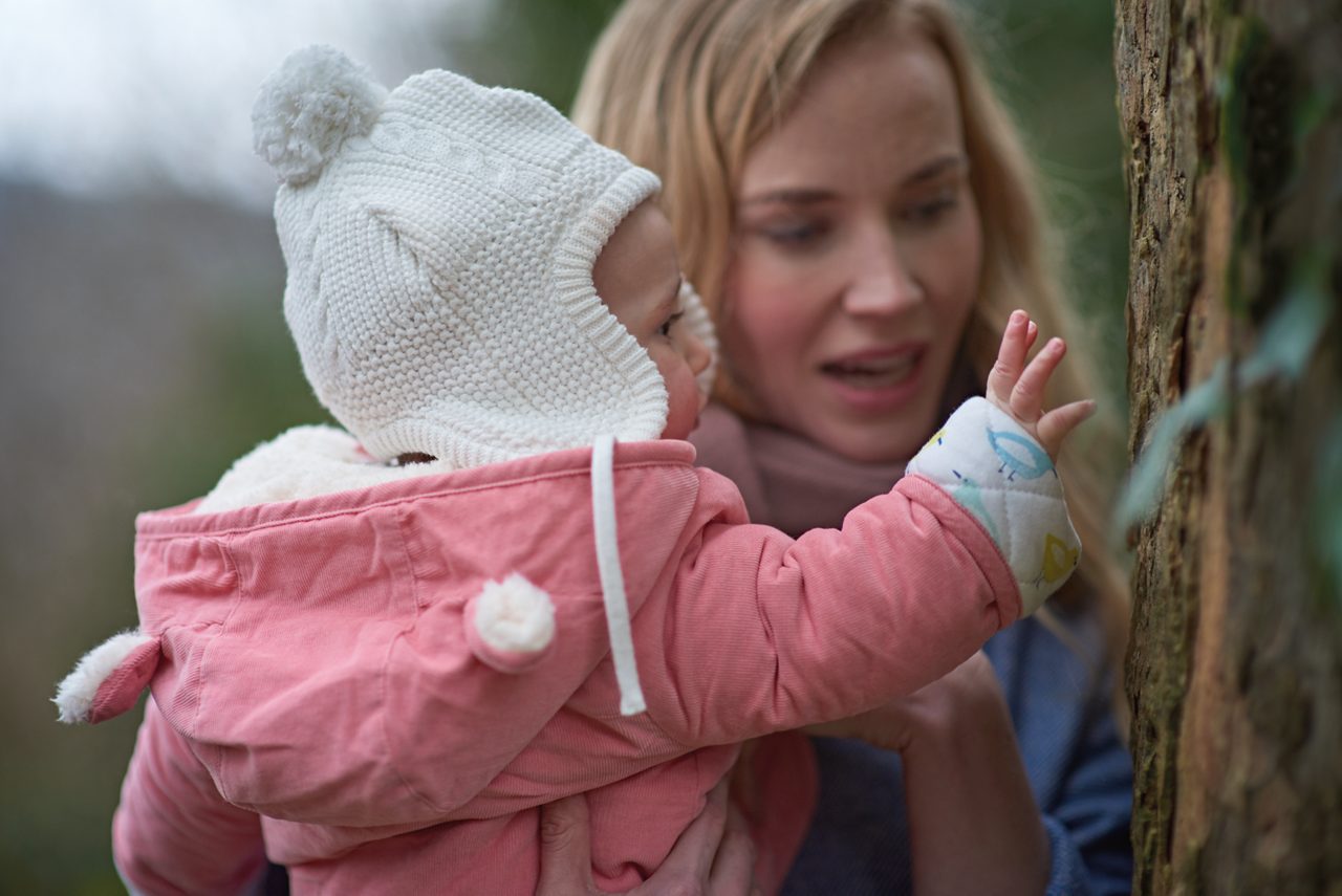 Baby auf Mamas Arm in einer rosa Winterjacke, mit einer weißen Wollmütze auf dem Kopf.