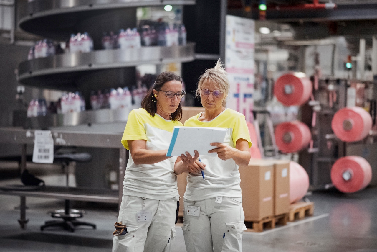 Zwei Frauen stehen in einem Werk und blicken auf einen Plan auf Papier