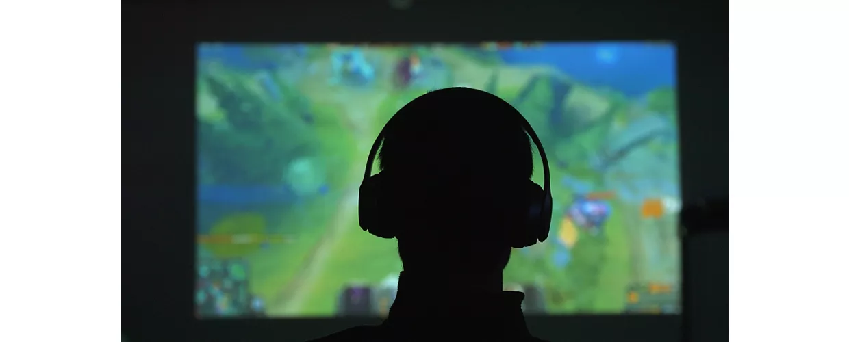 Silhouette di spalle di un uomo con delle cuffie che gioca con un videogame davanti a un grande schermo in una stanza scura