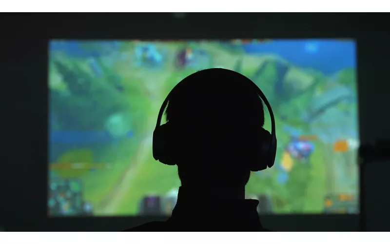 Silhouette di spalle di un uomo con delle cuffie che gioca con un videogame davanti a un grande schermo in una stanza scura
