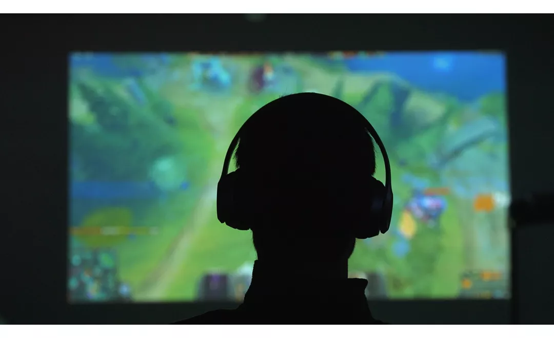 Silhouette di spalle di un uomo con delle cuffie che gioca con un videogame davanti a un grande schermo in una stanza scura